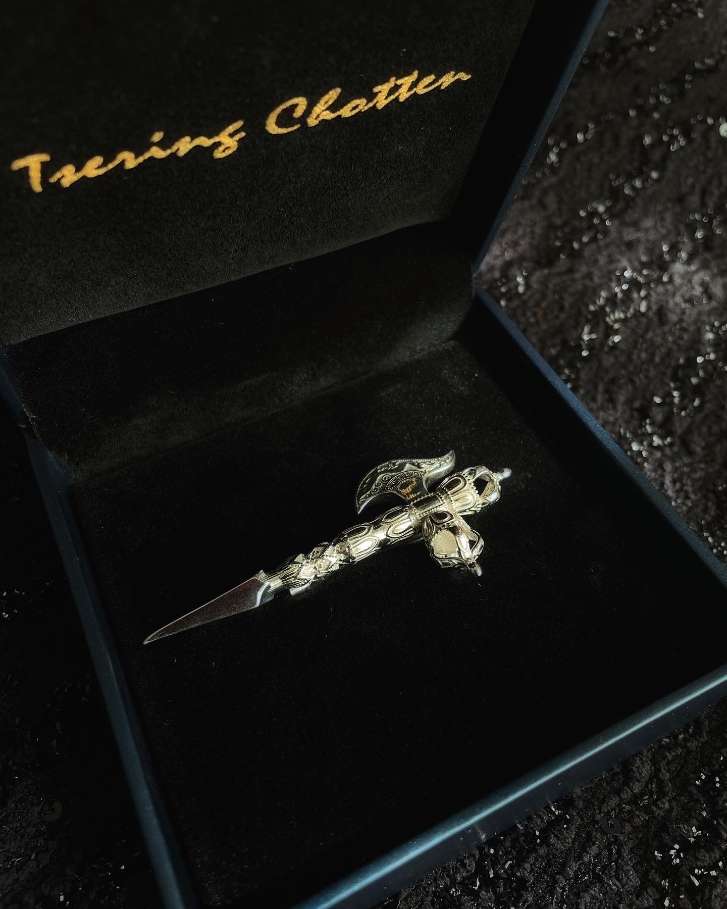 A silver colored brooch with a intricate design, displayed on a black surface with a velvet-lined box in the background. it is inspired by the dorjee symbol or vajra
