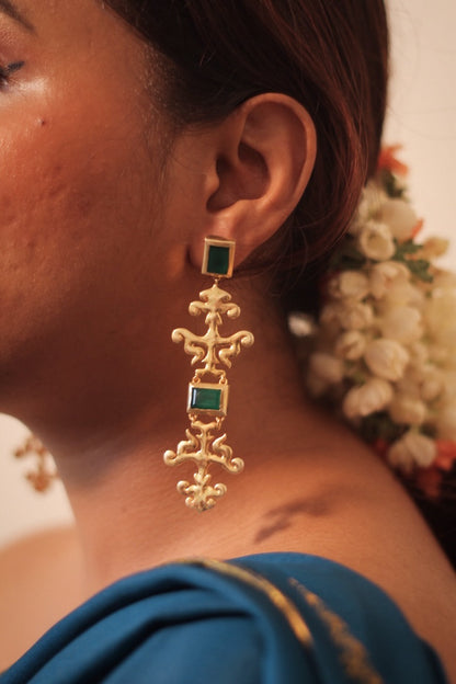 A close-up image of a woman's ear wearing a gold plated sterling silver art nouveau style dangling earring with a green square stone.